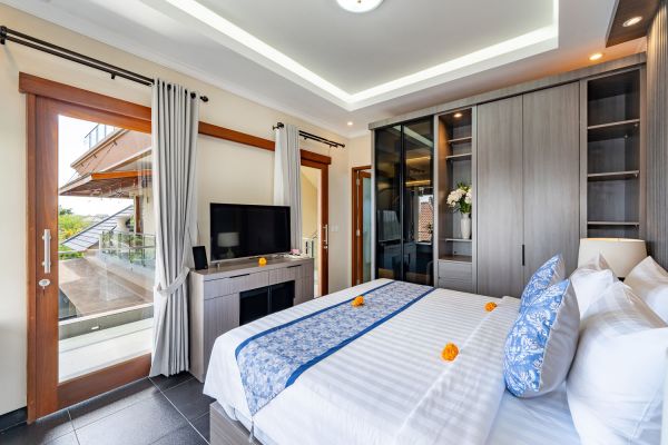 Luxurious Balinese villa bedroom with king bed, blue runner, open wooden doors to infinity pool, TV, and tropical decor.